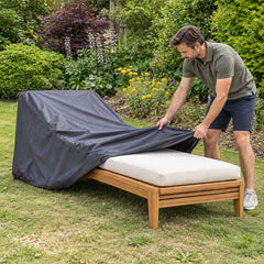 A man pulling a fitted charcoal chaise lounge cover over a cushioned teak sun lounger on a garden lawn