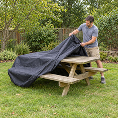 A man pulling a charcoal picnic table cover over a wooden picnic bench on a garden lawn, the cover going over the tabletop and both benches