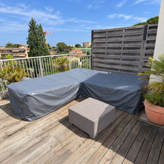 A grey made-to-measure cover fitted over a rattan corner sofa on a sunny balcony deck, with the rattan footstool beside it