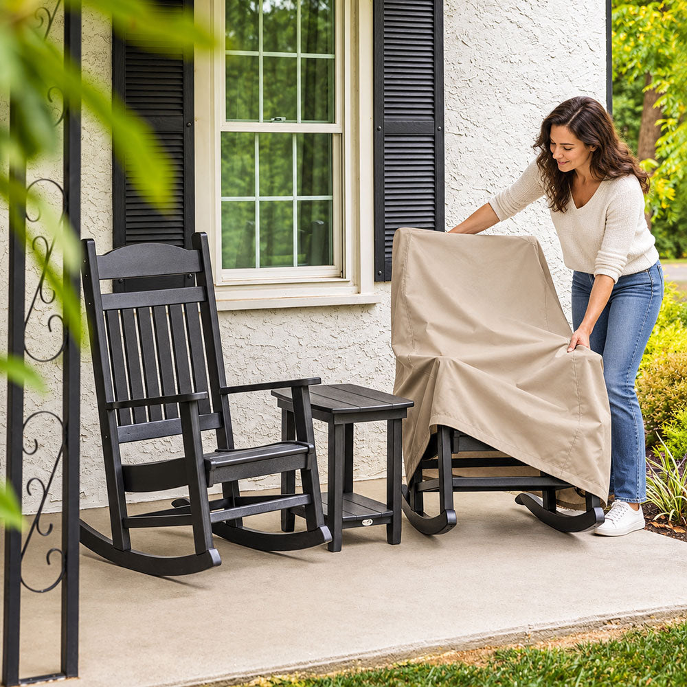 A woman pulling a beige rocking chair cover over a black rocking chair on a front porch, beside a matching uncovered rocking chair and side table