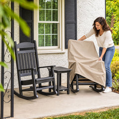 A woman pulling a beige rocking chair cover over a black rocking chair on a front porch, beside a matching uncovered rocking chair and side table